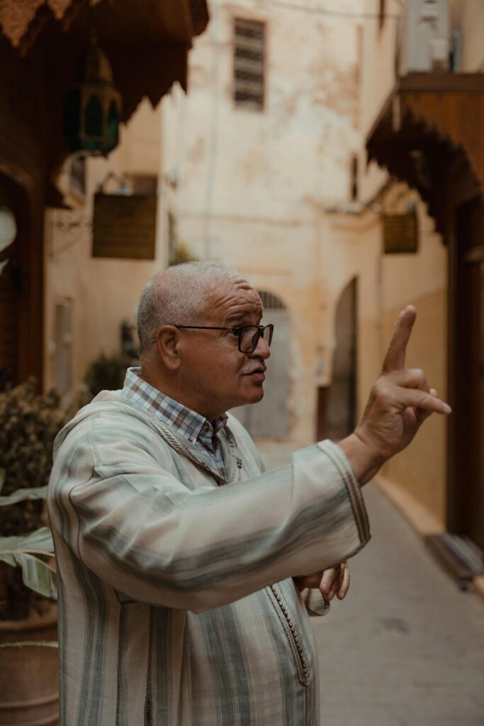 An adult man with glasses gestures while speaking in a historic street.